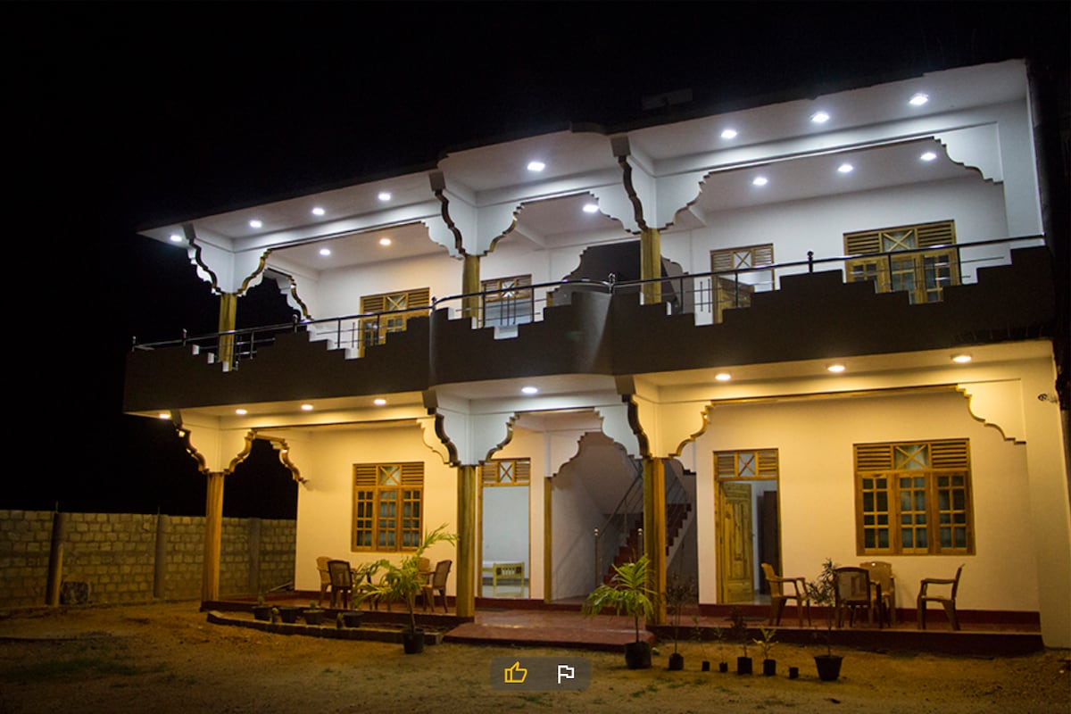 The exterior of a two-story building is illuminated against the night sky, showcasing detailed architectural features. Various outdoor seating areas are located on the patio, surrounded by lush greenery. Multiple windows allow natural light to enhance the welcoming atmosphere of the structure.