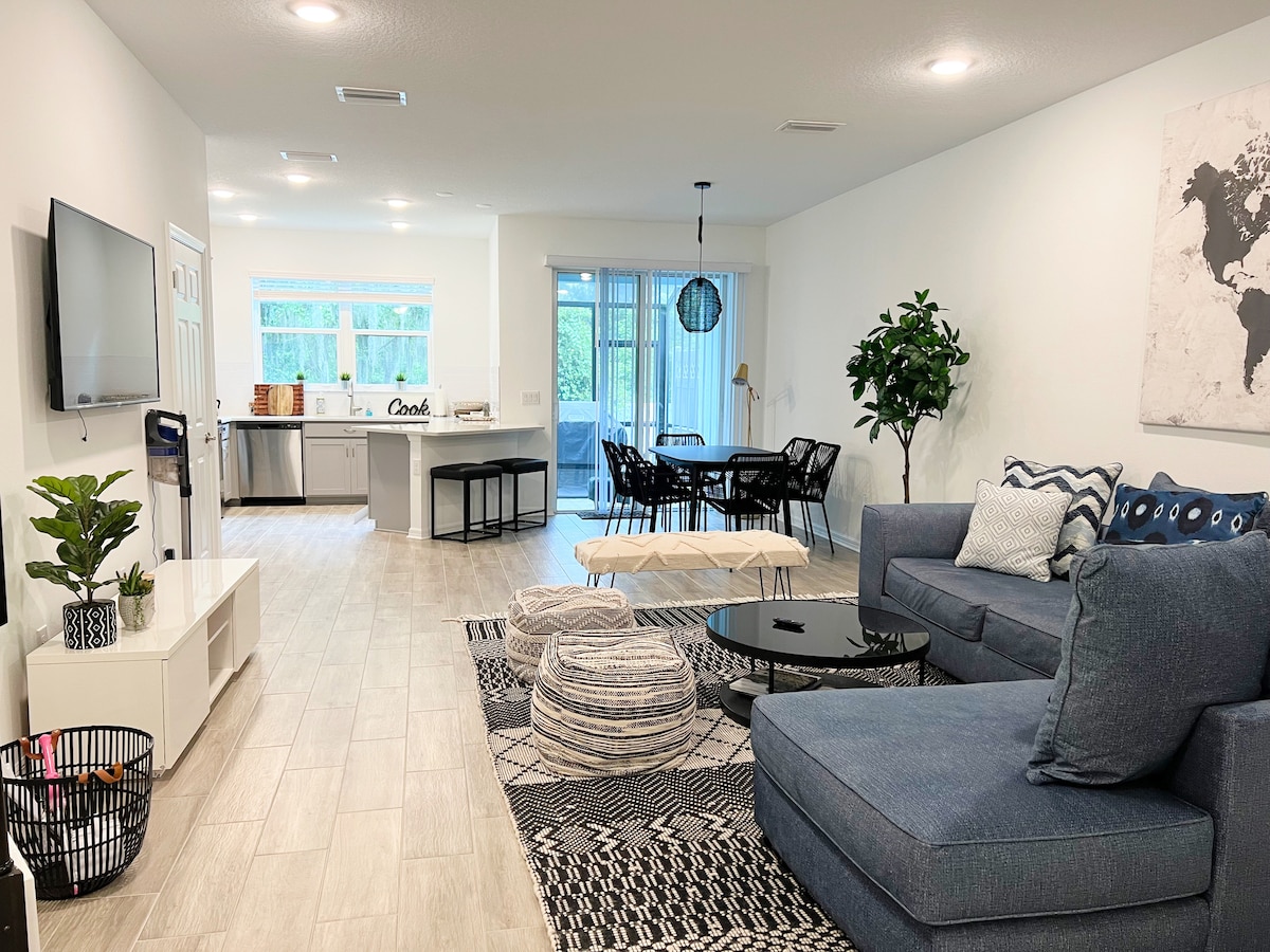 An open-concept living space features a comfortable sectional sofa with colorful cushions, a modern coffee table, and patterned area rugs. A light-filled kitchen and dining area are visible in the background, with a view of sliding glass doors leading to an outdoor space.