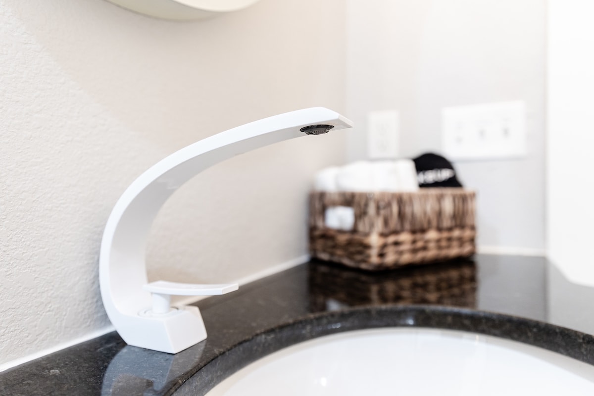 A modern faucet is positioned above a round sink with a sleek black countertop. Behind the faucet, a woven basket holds neatly rolled towels, contributing to the organized and tidy appearance of the bathroom space.