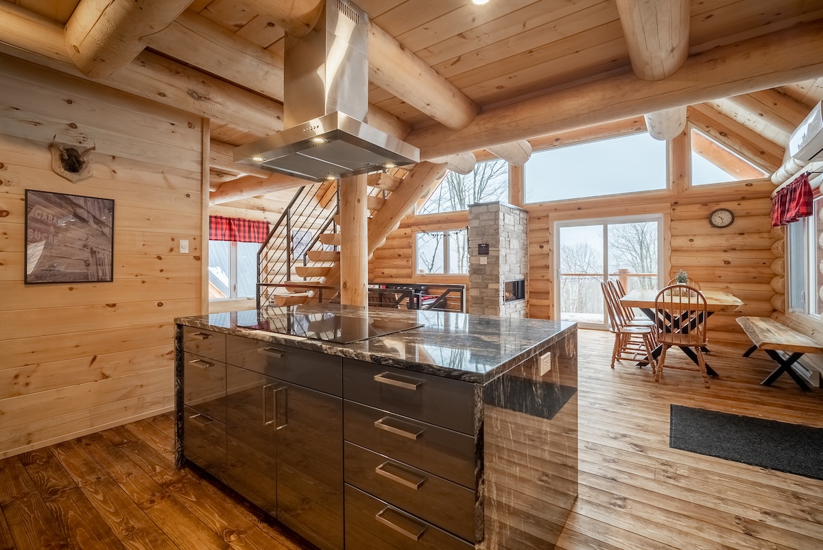 A kitchen area showcases a large central island with a dark surface and sleek storage drawers. Natural wood walls and beams create a warm ambiance, illuminated by overhead lighting. Nearby, a dining area with wooden chairs and a table is visible through large windows offering views of the outdoors.