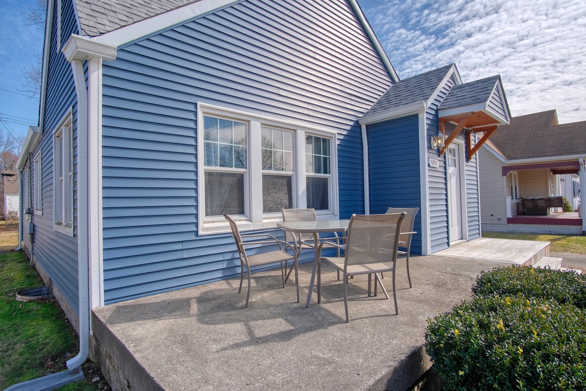 A blue exterior of the house is complemented by a welcoming porch area featuring a rectangular table surrounded by four chairs. Two large windows allow natural light to illuminate the space. A manicured hedge borders the porch, enhancing the appealing entrance.