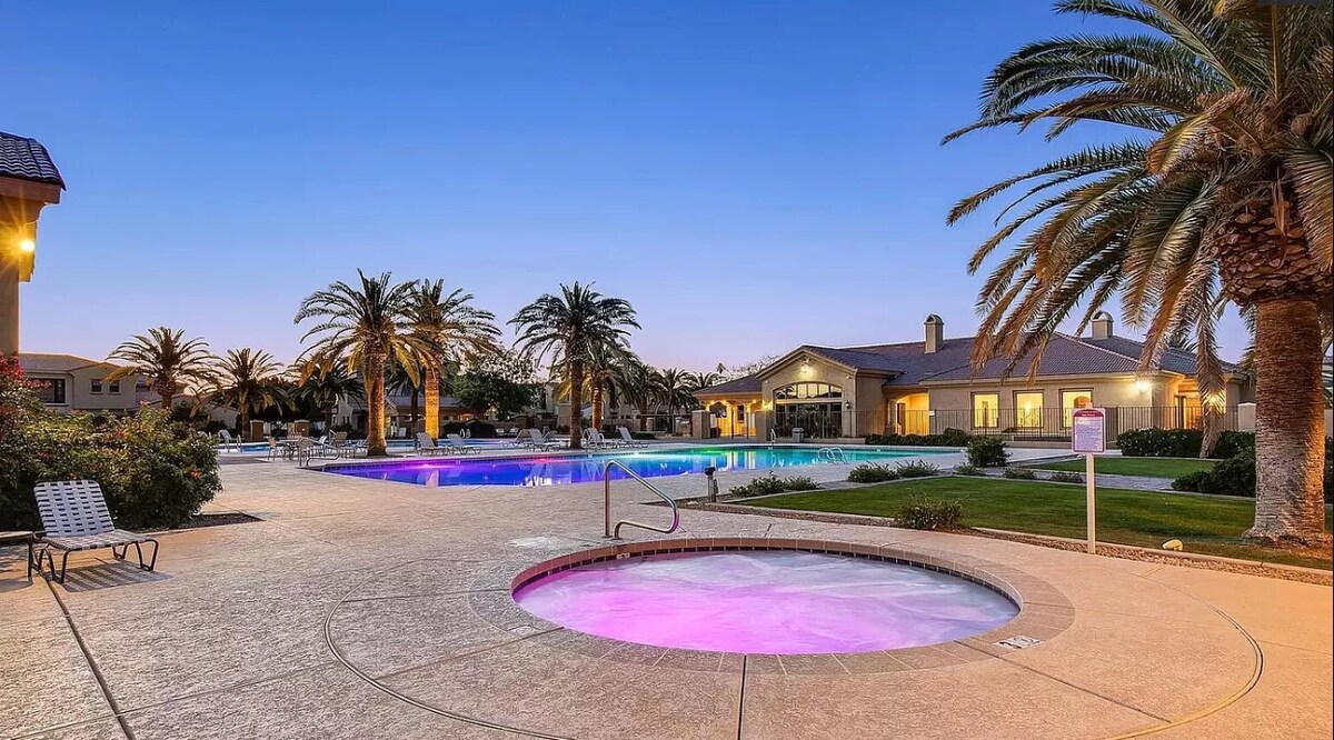 The resort-like area features two expansive Olympic-sized pools, one with purple lighting, surrounded by lush palm trees. A hot tub is positioned nearby, with well-maintained landscaping visible in the background. The clubhouse appears inviting, enhancing the community's recreational atmosphere.