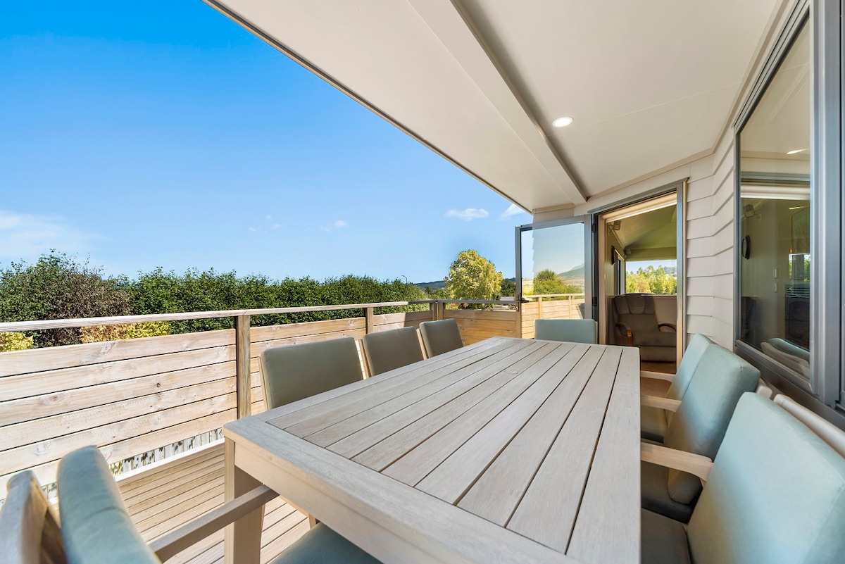 An outdoor dining area features a large wooden table surrounded by cushioned chairs. The space is enhanced by sliding glass doors that open to scenic views of greenery and a clear blue sky.