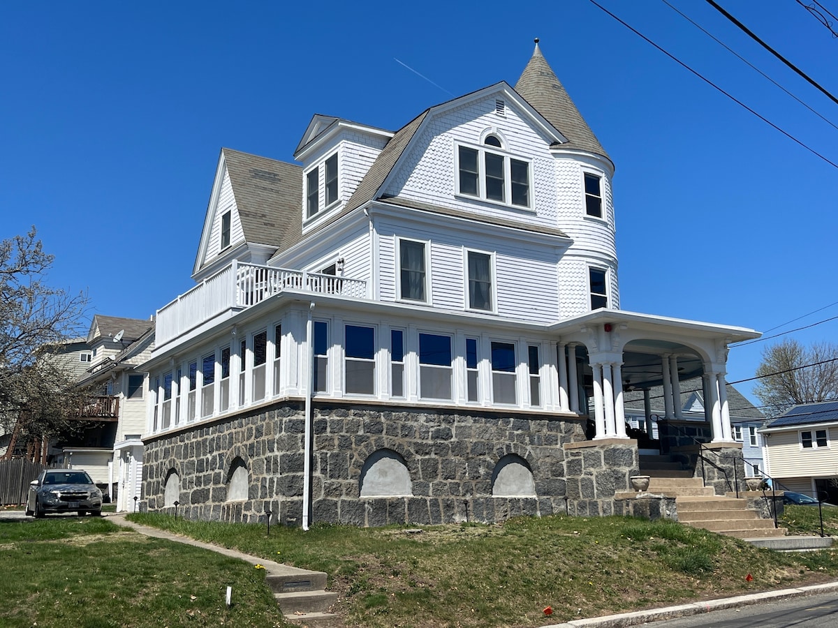 A grand Victorian home built in the 1880s is captured from a street view, showcasing its distinctive architecture and inviting wrap-around porch. Expansive windows reflect natural light, highlighting the building's historic charm and meticulously maintained exterior.
