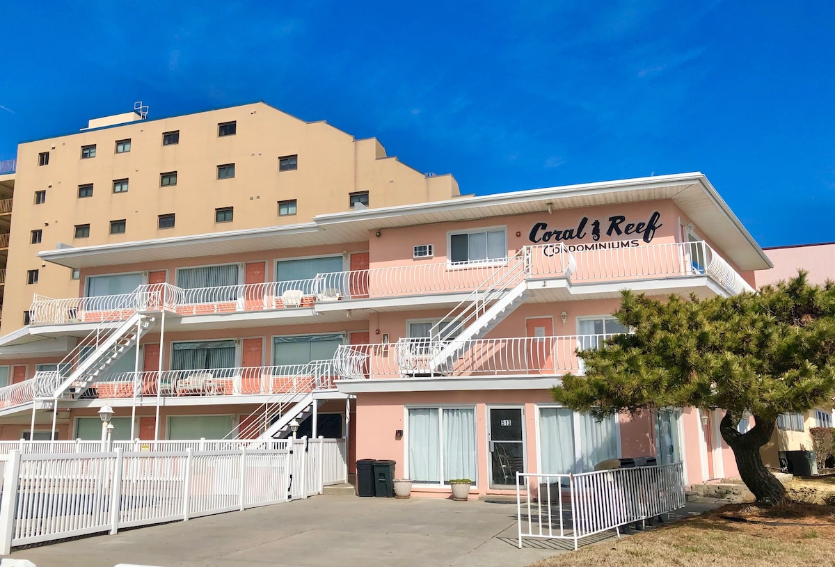 The exterior of a beachfront condominium complex is displayed, featuring a pale pink facade and white railings. Multiple staircases lead to the upper levels, while a neatly maintained courtyard area is framed by a low fence and greenery, under a clear blue sky.