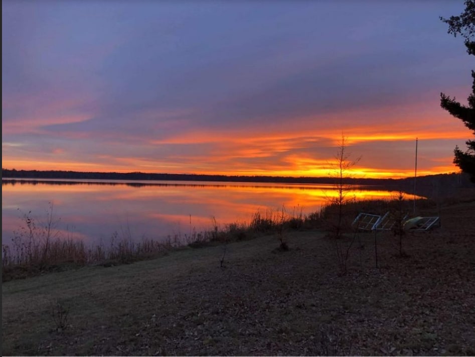 A serene lakeside view captures the vibrant colors of a sunset reflecting on the calm water. The grassy shoreline is bordered by tall grasses and scattered trees, creating a natural frame for the picturesque scene.