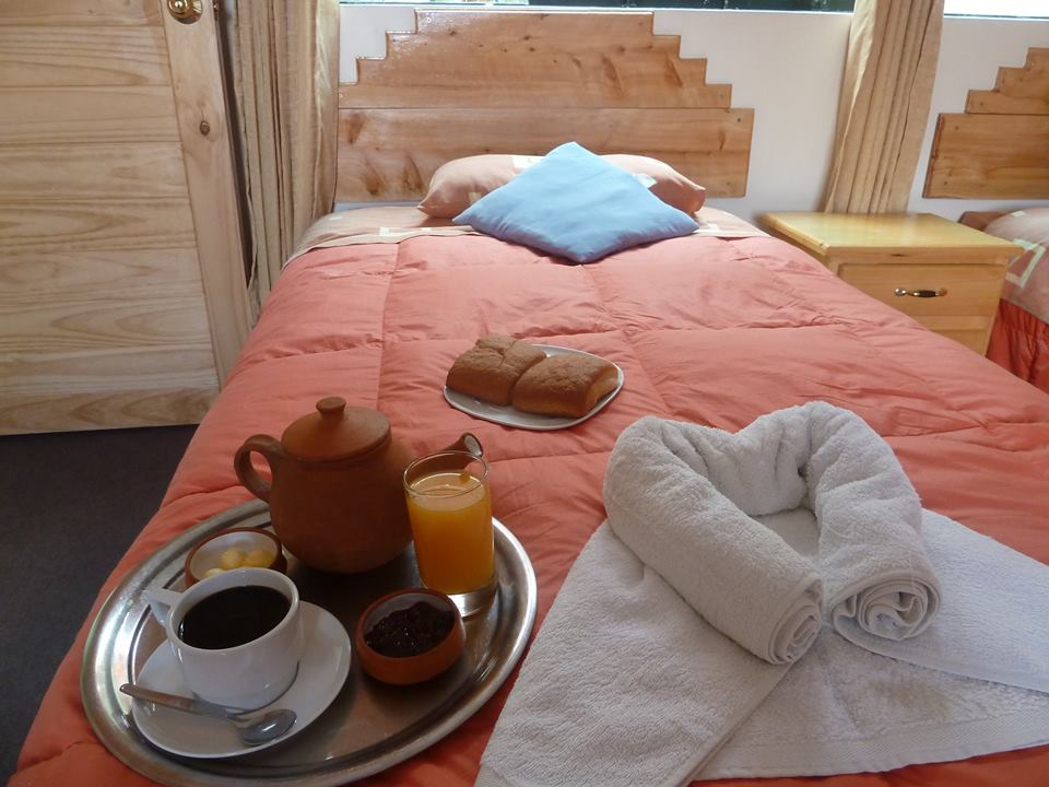 A cozy sleeping area is highlighted by a spacious bed dressed in soft orange bedding. A wooden bedside table holds a tray with coffee, juice, pastries, and condiments. Fresh towels are neatly arranged beside the bed, enhancing the welcoming setting.