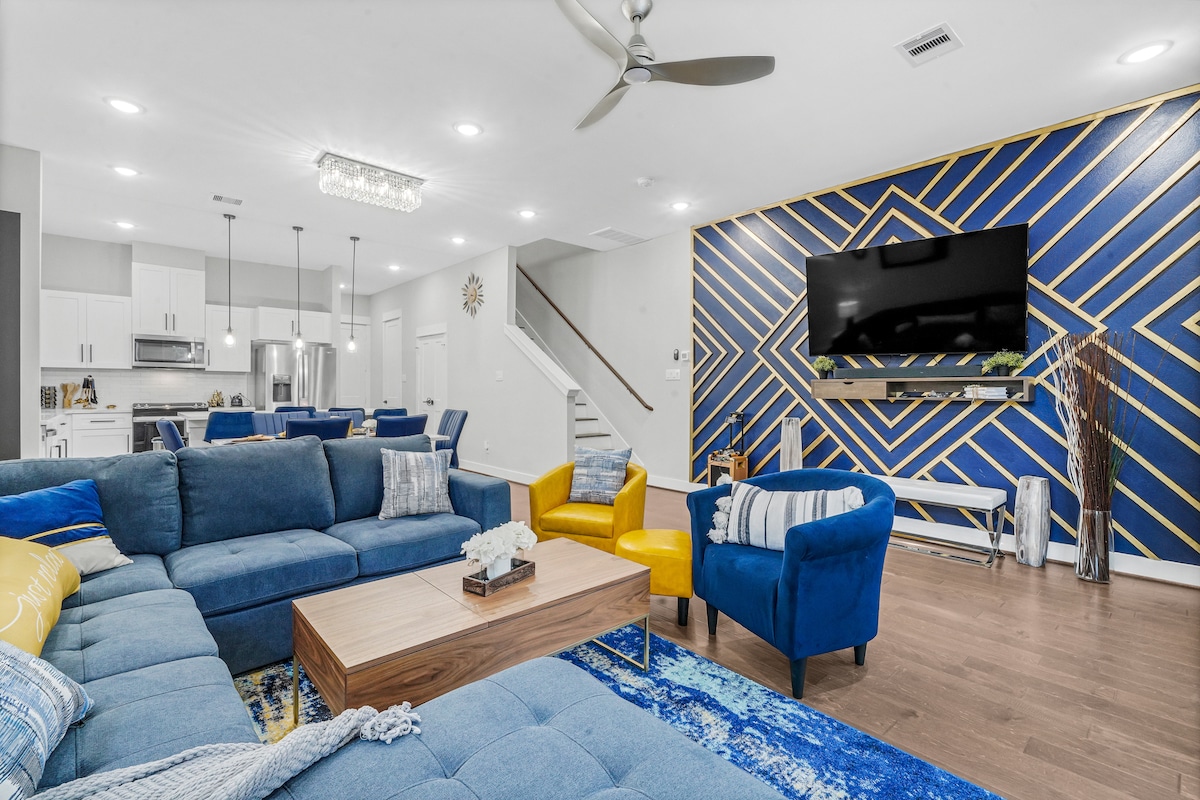 A spacious living area is furnished with a large blue sectional couch and a wooden coffee table. A vibrant blue and gold accent wall features a geometric pattern, while a flat-screen TV is mounted for viewing. Natural light fills the room, enhancing the modern decor.