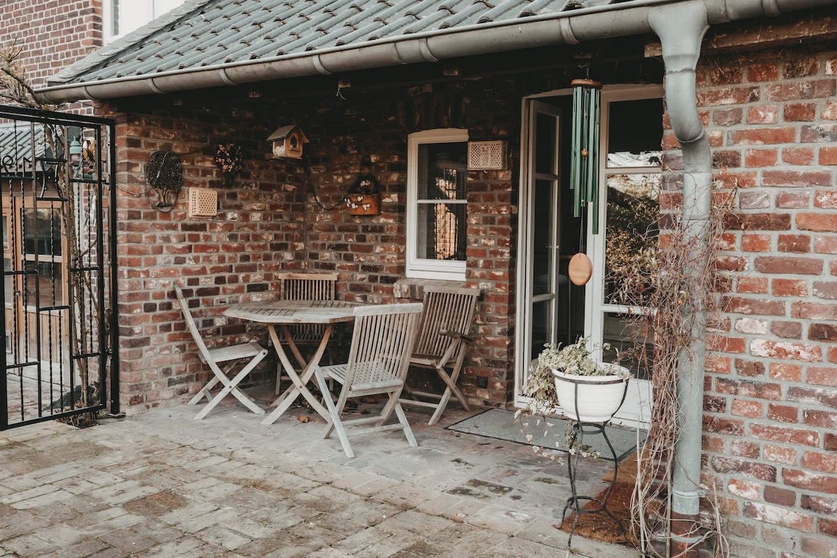 An outdoor terrace is framed by rustic brick walls, featuring a wooden dining table surrounded by folding chairs. A wind chime hangs near the door, and a potted plant is placed on the ground. The entrance leads to additional indoor space.
