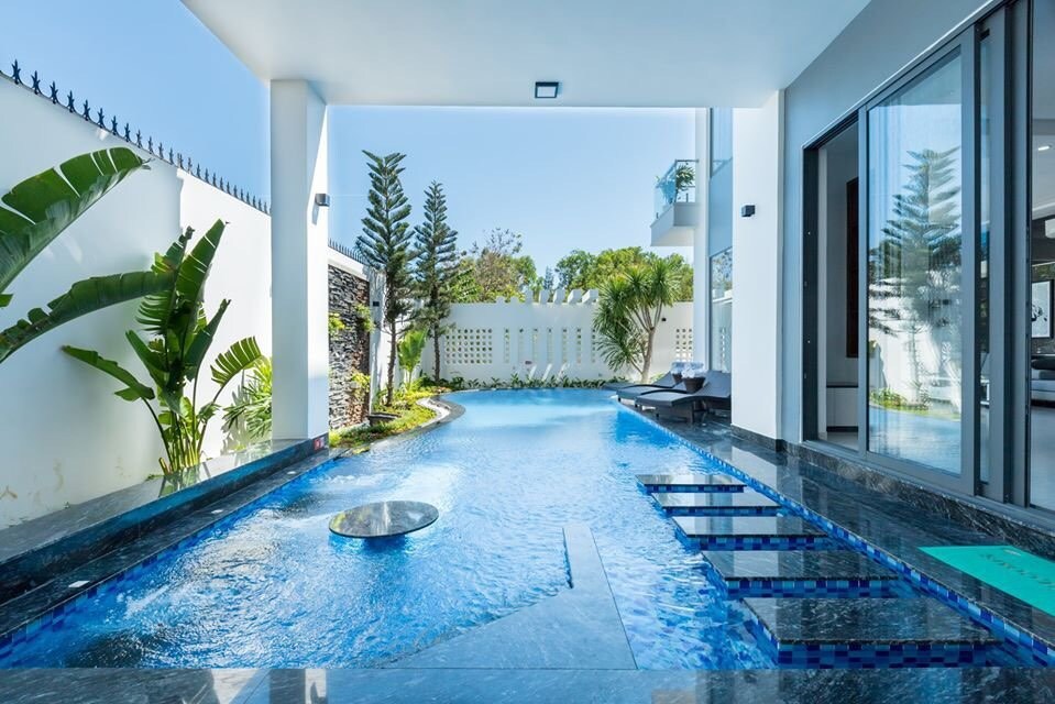 A modern swimming pool is featured in the outdoor area, surrounded by lush greenery. The pool's clear blue water reflects the sunlight, and a smooth surface creates a seamless transition to the villa's interior. Comfortable seating areas are positioned nearby for relaxation.