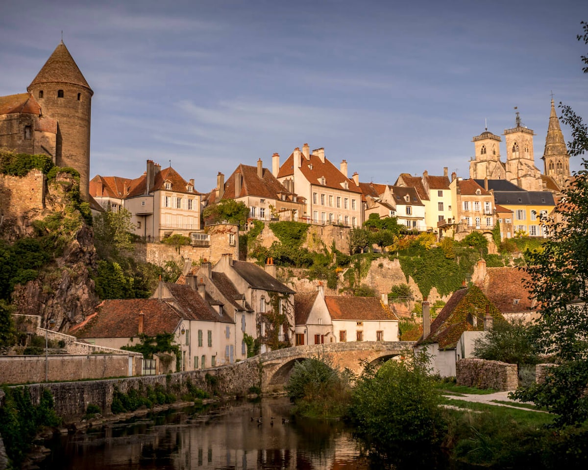 A scenic view of Semur-en-Auxois showcases the medieval architecture perched upon a hillside. The stone towers and charming buildings are framed by lush greenery, along with a peaceful river reflecting the surrounding landscape. A historic bridge spans the water, adding to the picturesque setting.
