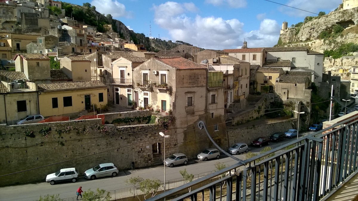A view from the balcony captures a scenic landscape of Modica, featuring a mix of historical buildings and residential homes. The image shows a street lined with parked vehicles and lush greenery along a stone wall, with hills rising in the background under a partly cloudy sky.