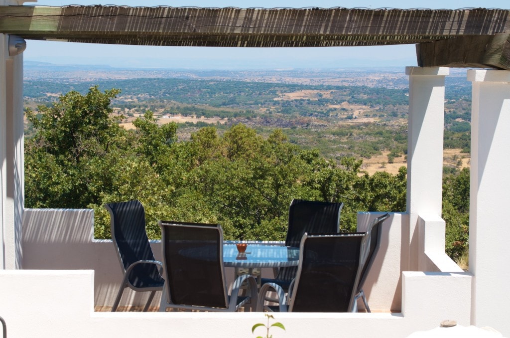 A terrace features a table surrounded by four chairs, offering an unobstructed view of the valley and landscape beyond. The space is shaded by a wooden pergola, with white walls framing the scene, creating a serene outdoor dining area.