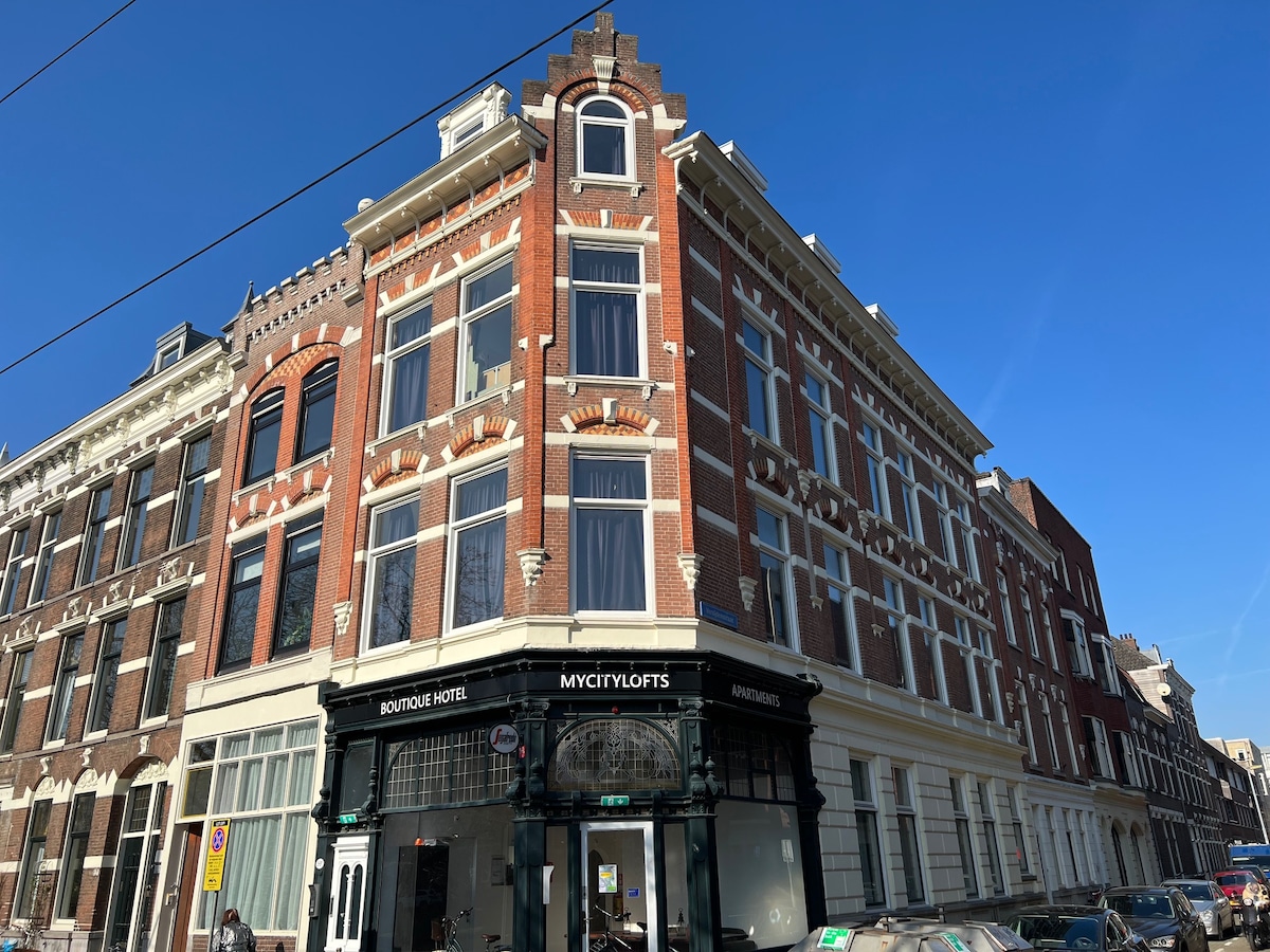 The exterior of a red brick building is showcased, displaying intricate architectural details. Large windows reflect natural light, and the main entrance features a stylish sign indicating a boutique hotel. A clear blue sky serves as the backdrop.