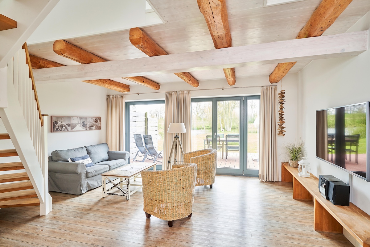 The living area features exposed wooden beams and large glass doors that provide access to an outdoor space. Comfortable seating arrangements are visible with a light-colored sofa and two woven chairs. Natural light fills the room, complemented by neutral-colored curtains.