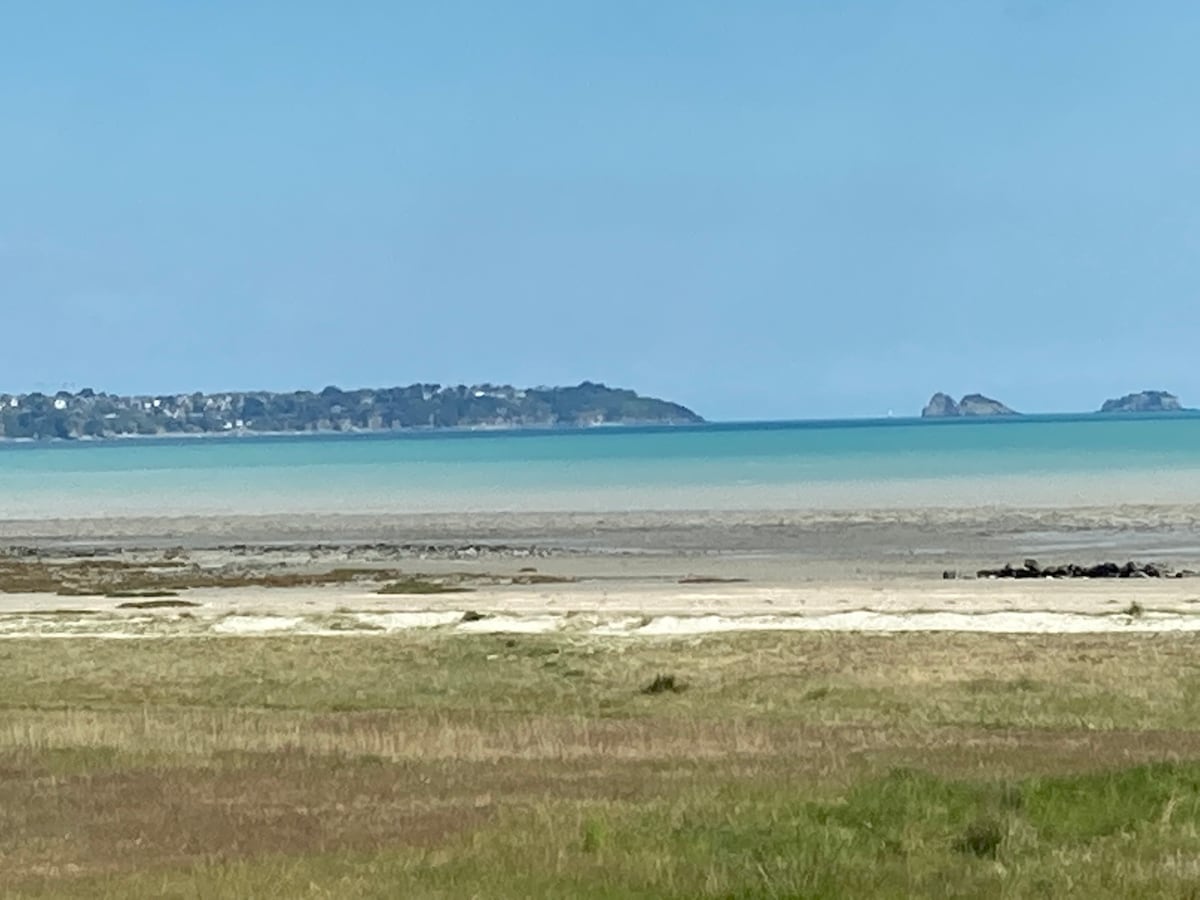 A serene view of the coastline, featuring calm turquoise waters gently merging with the sandy beach. A distant green landscape is visible along the shoreline, with subtle outlines of hills and rocky formations on the horizon.