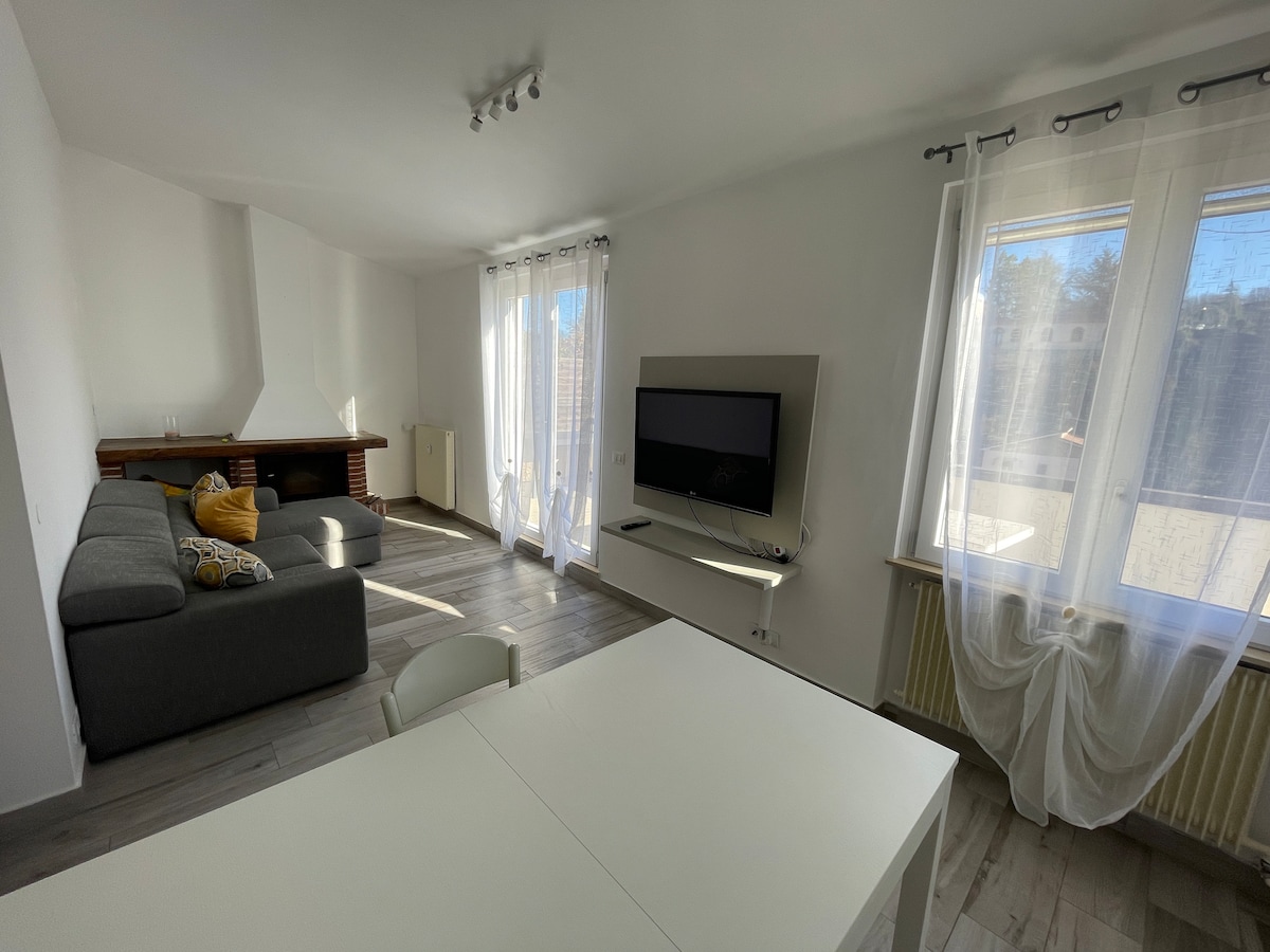 A bright living area features a spacious gray sectional sofa, complemented by a yellow decorative pillow. Large windows invite natural light, while a flat-screen TV is mounted on the wall. A dining table is positioned nearby, creating an open and welcoming atmosphere.