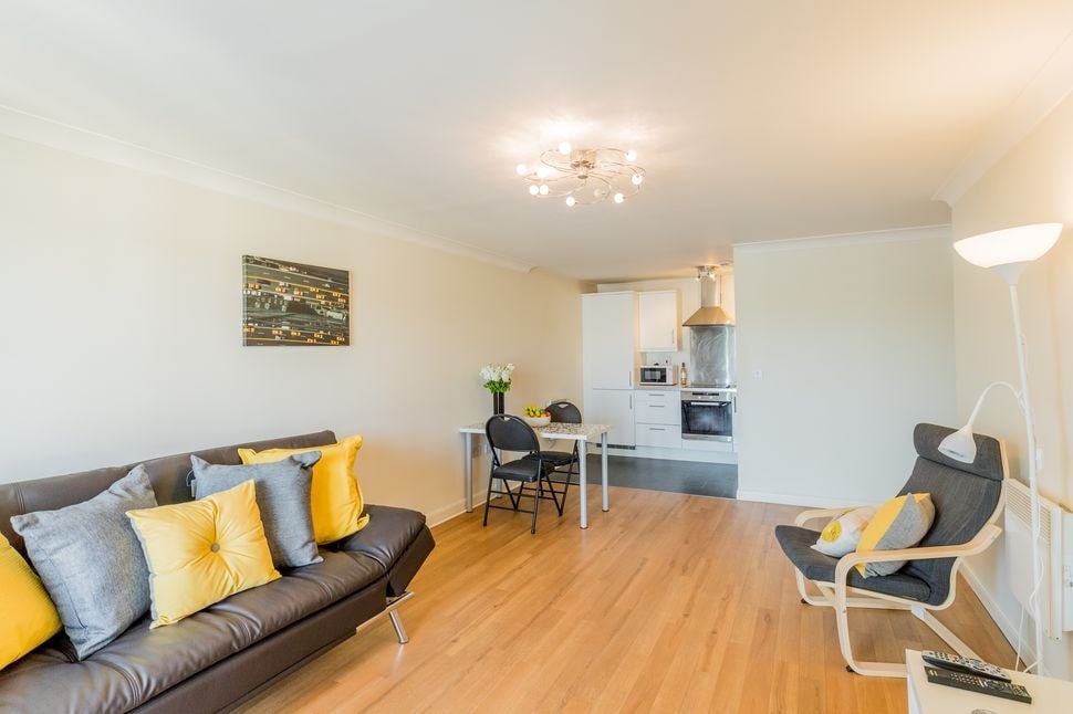 A living area features a dark sofa adorned with yellow and grey cushions, paired with a light chair. A dining table with two chairs is visible in the background near a modern kitchen setup. Soft lighting highlights the room's neutral walls and wooden flooring.