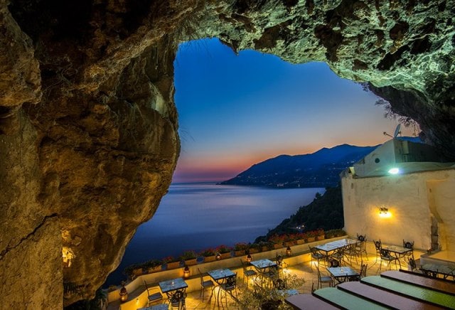 A natural cave entrance showcases a stunning view of the coastline at sunset, with soft hues of orange and blue in the sky. Outdoor dining tables and chairs are arranged in a terrace setting, complemented by vibrant flower pots along the edges.