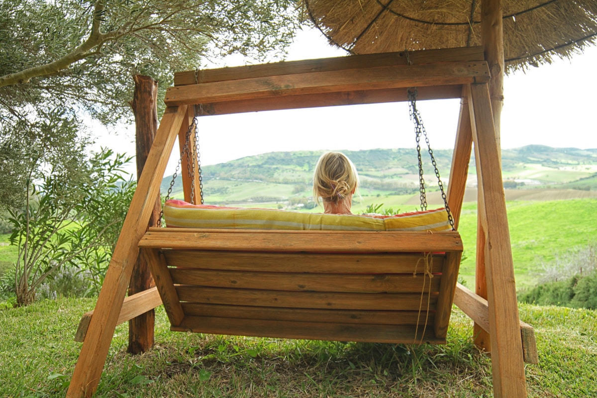 A wooden swing is positioned beneath a straw-covered shelter, overlooking lush green hills and valleys. The swing is securely suspended by chains, providing a serene spot to enjoy the expansive views of the surrounding landscape.
