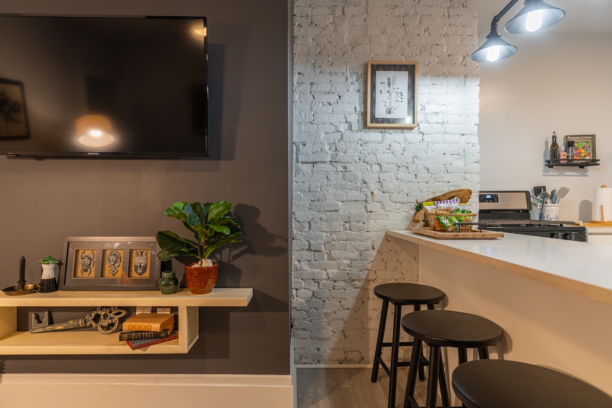 A modern living space features a mounted TV on a dark wall, with a minimalist shelf displaying framed art and a potted plant. An open kitchen area is seen with a counter and three black stools, illuminated by overhead lights, and a light-colored brick wall adds texture.