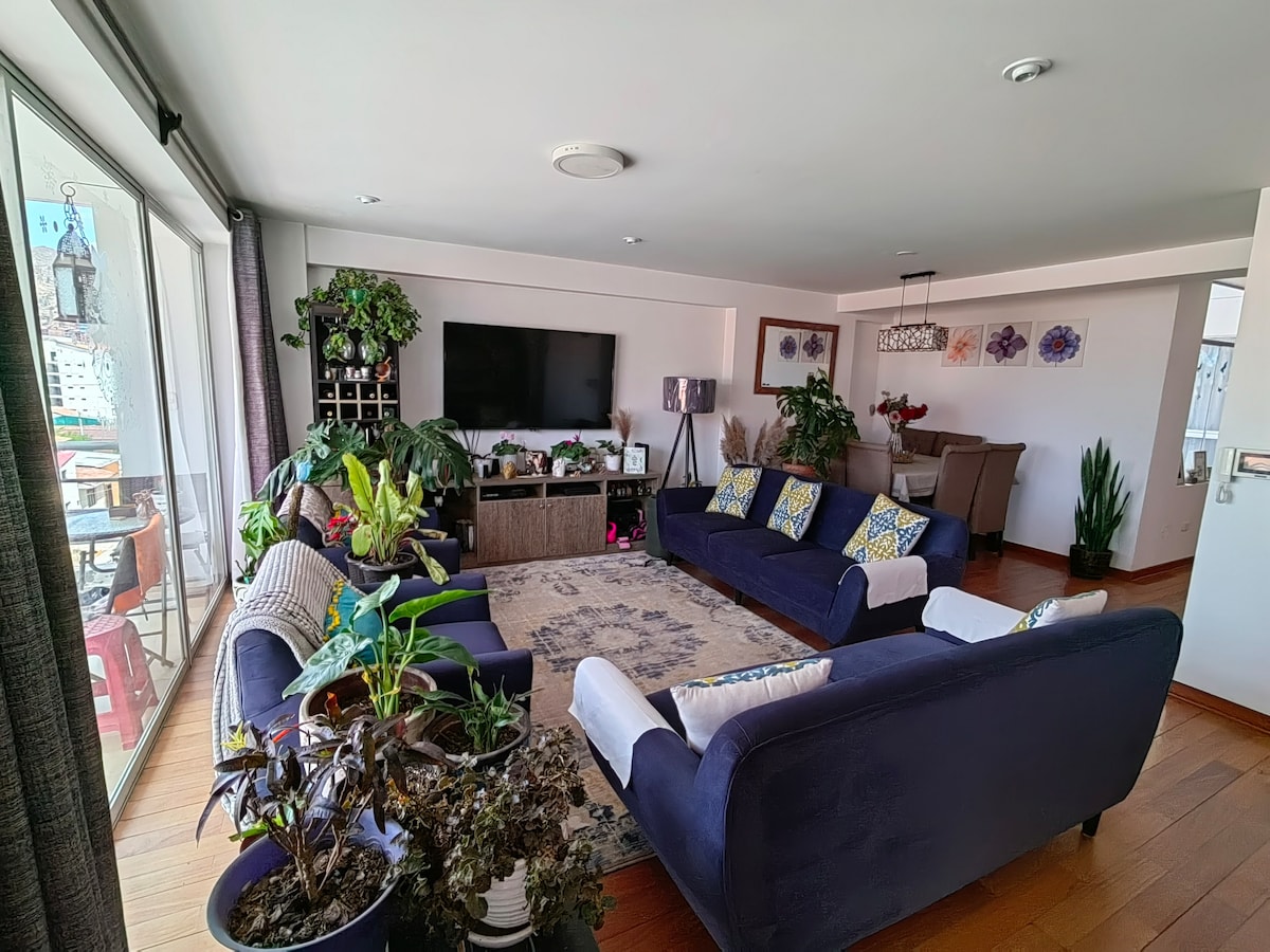 A spacious living area features a large, plush sofa arranged around a central decorative rug. Multiple plants add greenery, while a television is mounted on the wall. Large windows provide natural light and access to a balcony with outdoor seating.