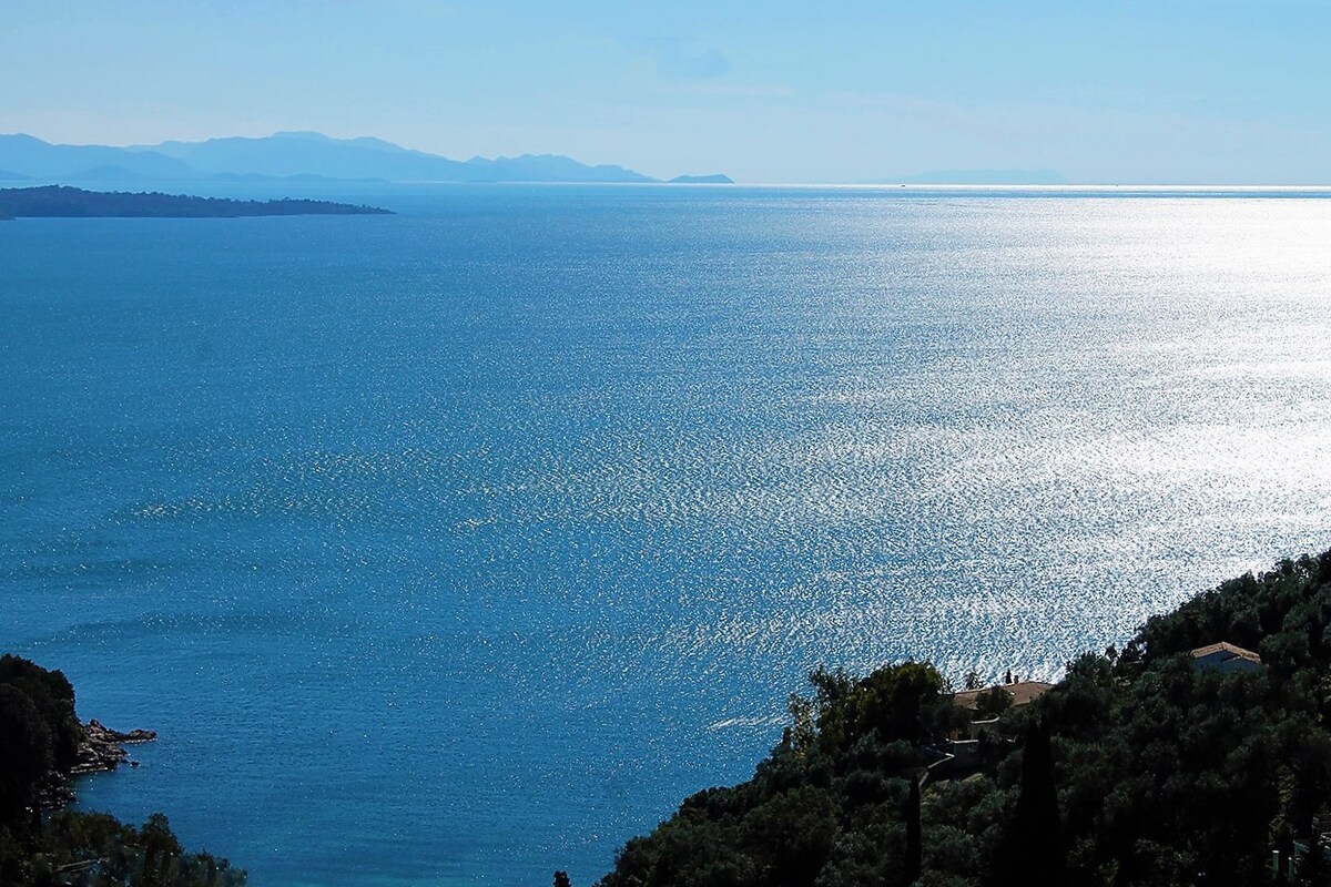 A panoramic view of the shimmering blue sea extends towards distant mountains under clear skies. The coastline is softly framed by lush greenery, with hints of distant land visible on the horizon, reflecting the sunlight across the water's surface.