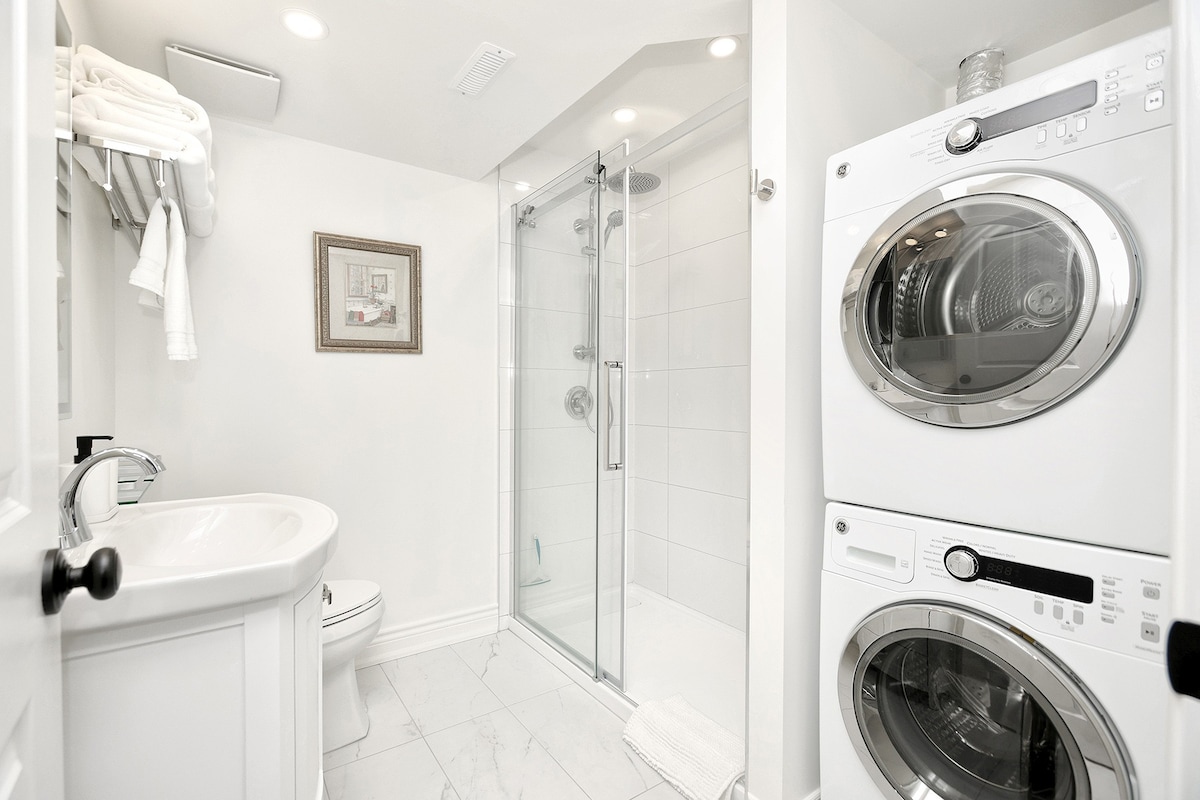 The bathroom features a glass-enclosed shower with a rain shower head, complemented by a large wall-mounted washer and dryer. A small wall-mounted shelf holds fresh towels, while a framed piece of art adds a decorative touch. The layout is bright and functional.