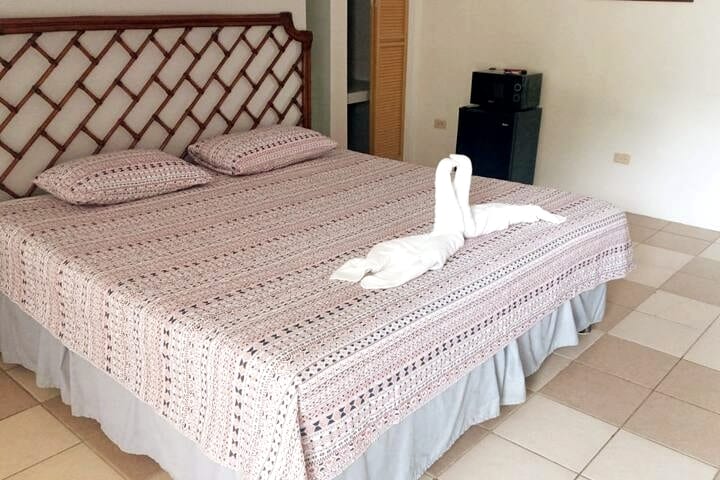 A spacious bed with a patterned coverlet is present in the room. Two pillows are arranged at the head of the bed, with a decorative towel swan positioned in the center. A small refrigerator is visible in the background, contributing to the room's functionality.