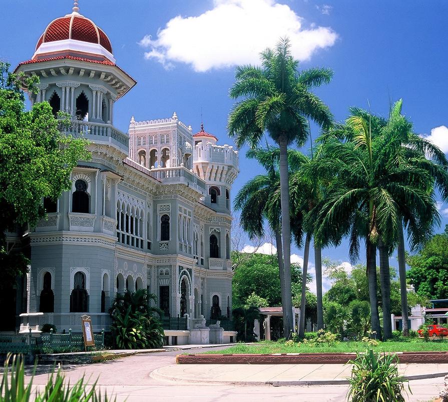 An elegant colonial building is surrounded by lush palm trees and greenery, showcasing intricate architectural details and vibrant colors under a clear blue sky. The spacious façade features a striking combination of domes and arches, creating a captivating visual appeal.