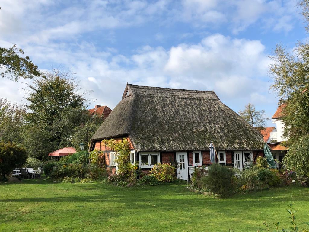 A charming thatched-roof house is surrounded by lush greenery and colorful flowering plants. The building features multiple windows framed with white shutters and is complemented by a well-maintained lawn. Nearby, outdoor seating areas under umbrellas are visible, inviting relaxation.