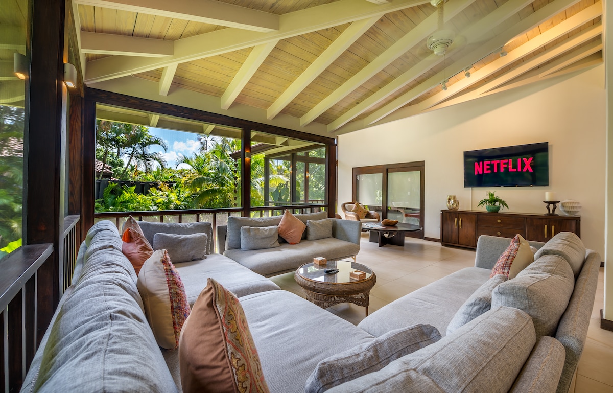 A spacious living area features a large sectional sofa adorned with colorful cushions. Natural light floods through expansive windows, revealing lush greenery outside. A Smart TV displays the Netflix logo, while a wooden coffee table sits in the center, inviting relaxation and entertainment.