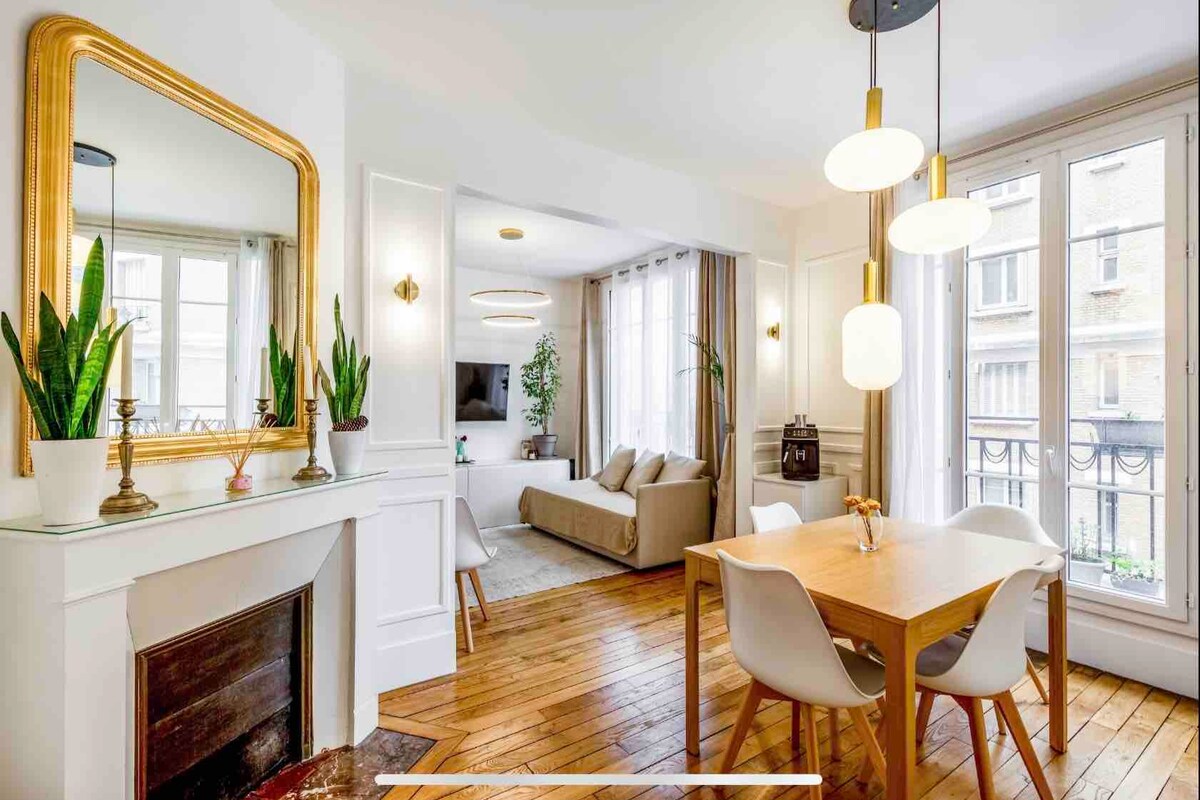 The dining area features a wooden table with white chairs, creating a bright space accented by natural light. A mirror with a gold frame adds elegance. A cozy living area, visible in the background, includes a sofa and a flat-screen TV beside large windows.