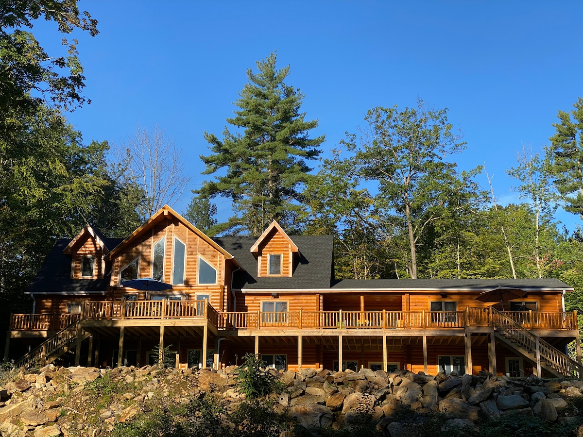 A newly built log home is set against a backdrop of tall trees and a clear blue sky. The two-story structure features a wraparound deck, large windows, and unique architectural details that enhance its rustic charm. A stone landscape surrounds the base of the home.