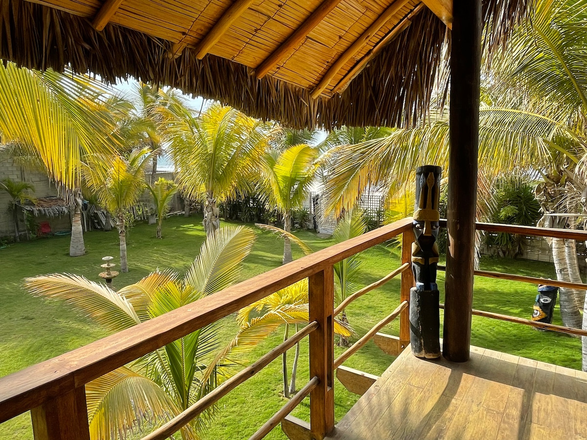 A wooden balcony overlooks a lush green garden filled with palm trees, creating a serene outdoor setting. The thatched roof provides shade, while the sunlight filters through the foliage, highlighting the vibrant colors of the landscape.
