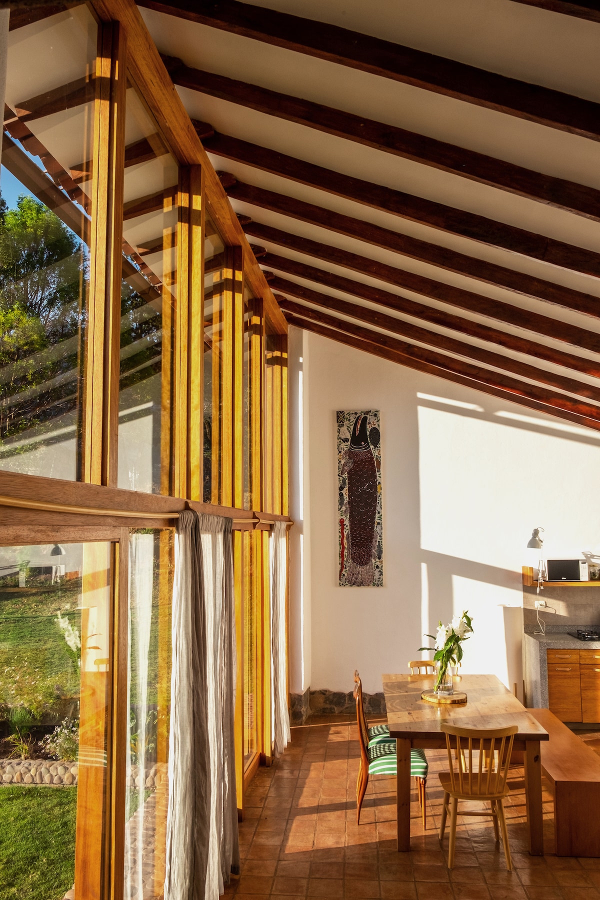 Natural light fills a spacious dining area, where large glass walls offer views of the surrounding garden. A wooden dining table is accompanied by chairs, and a piece of local artwork decorates the wall. The wooden beams of the sloped ceiling enhance the open feel.