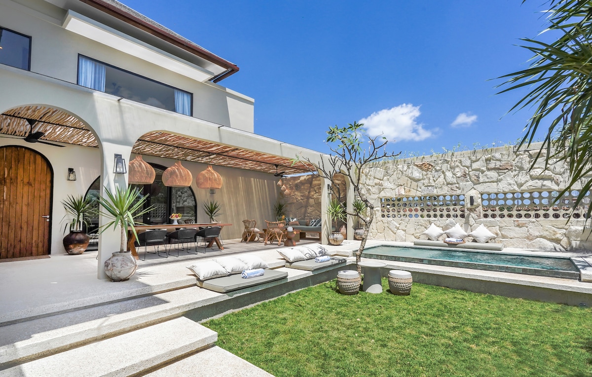 The outdoor space features a well-maintained garden with green grass and a private pool. A dining area with a large table sits under a covered terrace adorned with wooden light fixtures. Distinctive stone walls frame the scene, enhanced by tropical plants and decorative cushions.