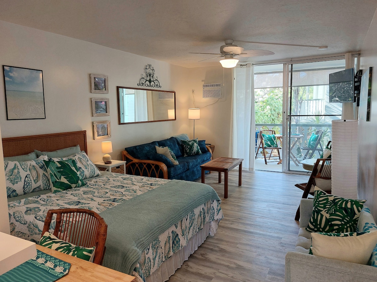 A bright studio space features a spacious bed adorned with tropical-themed bedding. Comfortable seating is provided near a large window, with natural light filtering through sheer curtains. A dining table is visible in the background, creating a cozy atmosphere. Decorative art pieces add character to the walls.