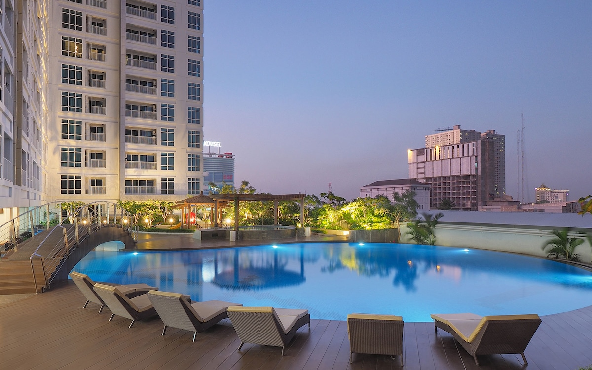 A large outdoor swimming pool is surrounded by a wooden deck and several lounge chairs. Lush greenery and palm trees are visible in the background, while the sky transitions from orange to deep blue as dusk approaches, highlighting the modern buildings of the city beyond.