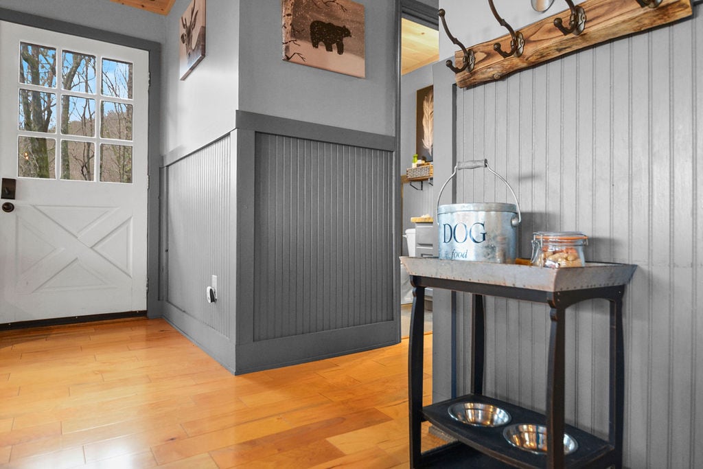 A welcoming entryway features a wooden console table with dog bowls and a metal container. A white door adorned with glass panes allows natural light to brighten the space. Gray paneling adds warmth to the setting, complemented by earth-toned wall art.