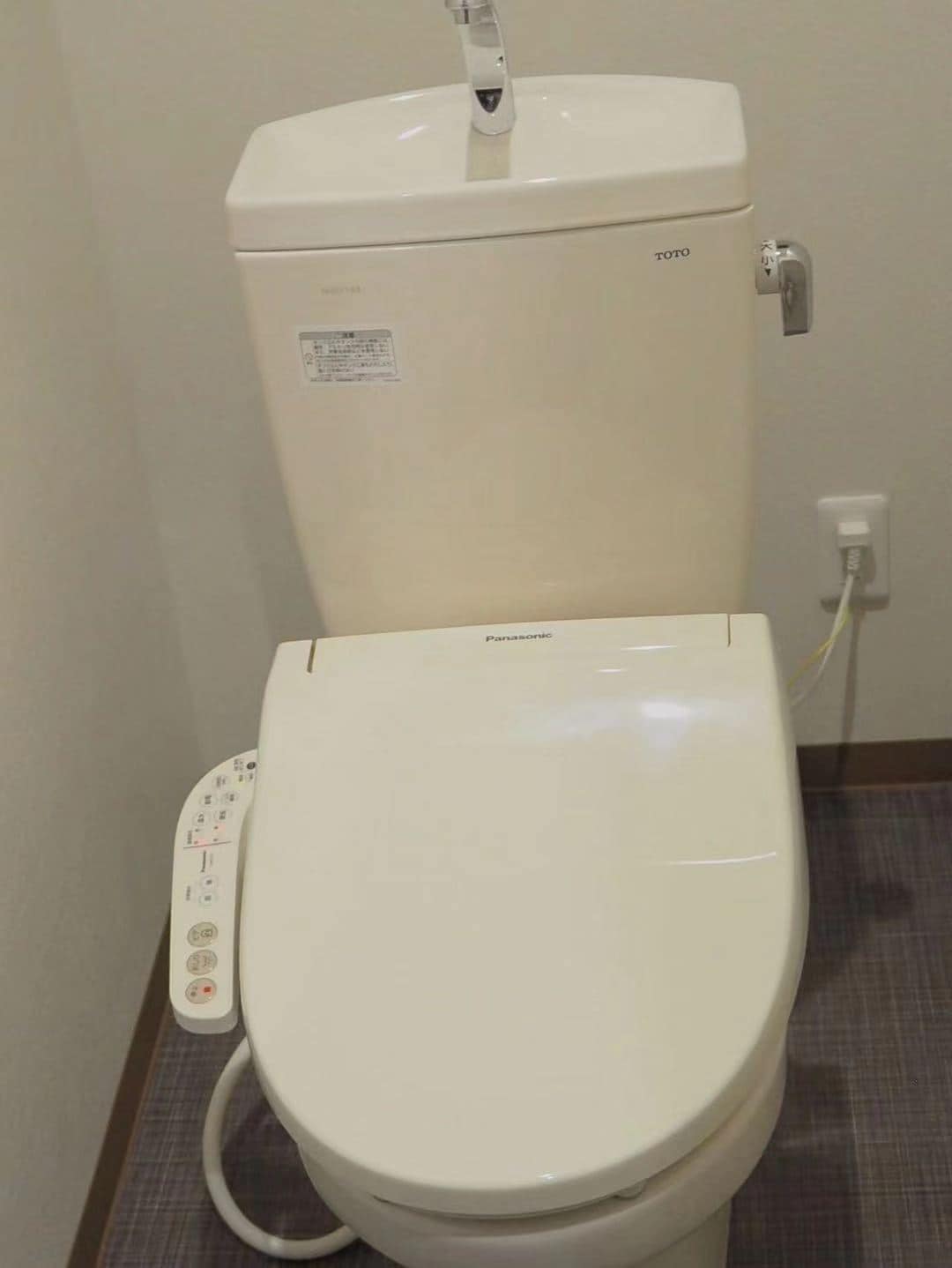 A modern toilet is positioned against a plain wall, featuring a sleek design in neutral tones. A control panel is attached to the side, indicating advanced functionality. A power outlet is visible nearby, ensuring accessibility to electrical needs.