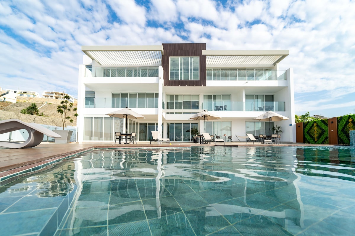 The modern building is shown from the perspective of the pool, highlighting its clean lines and large windows. Sun loungers and umbrellas are arranged around the pool area, which reflects the sky, creating a serene outdoor space. Lush greenery borders the property.