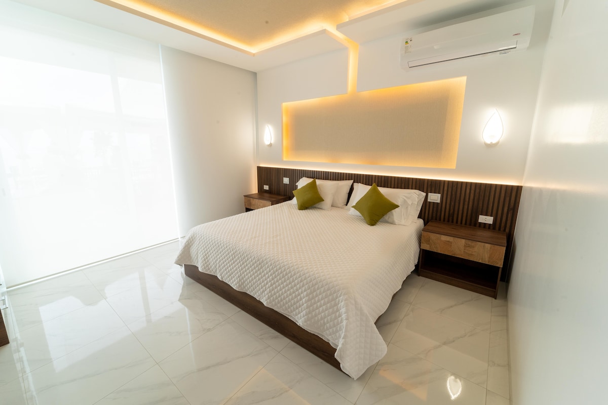 A spacious sleeping area is presented with a large bed adorned with white bedding and accent pillows. Soft lighting highlights the wall behind the bed, while two bedside tables complement the overall design. Natural light enters through expansive glass doors, creating a bright atmosphere.