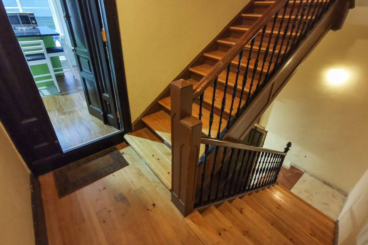 A well-lit staircase is visible, featuring polished wooden steps and a handrail. The entryway is observed at the top, leading into a kitchen area. The warm wood tones of the flooring enhance the welcoming feel of the space.