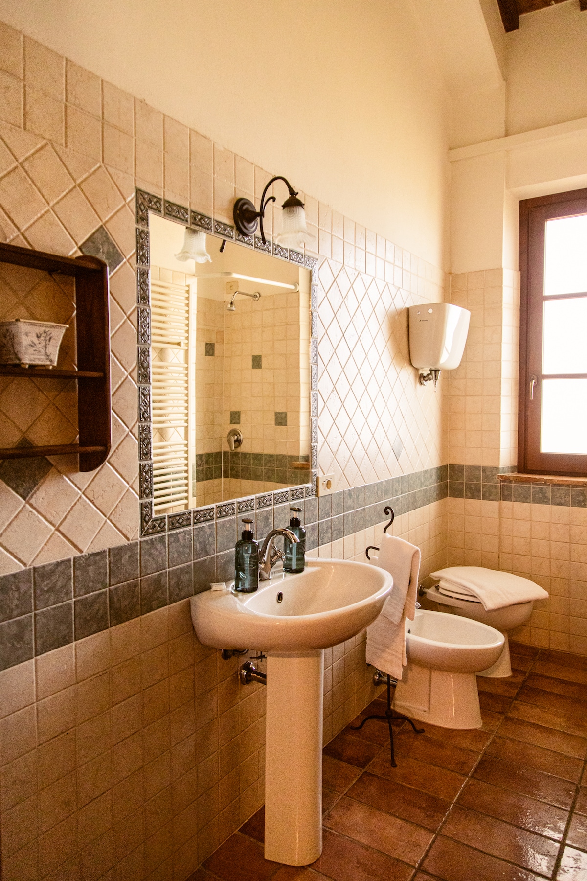 A well-appointed bathroom features a pedestal sink with modern faucets and toiletries. A mirror enhances the space, while a bidet and toilet are conveniently positioned nearby. Natural light enters through a window, accented by delicate wall tiles in muted tones.