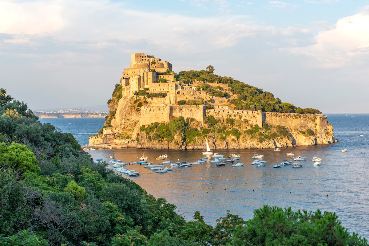An iconic castle is prominently situated atop a rocky island, surrounded by gentle waves and lush greenery. Small boats are anchored nearby, reflecting a serene coastal scene under a bright sky, showcasing a picturesque view of the historical landscape.
