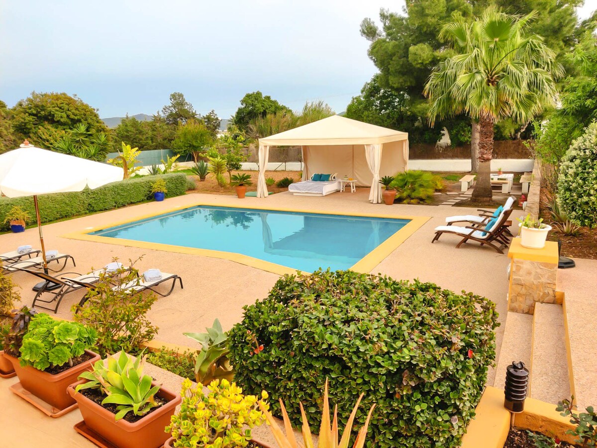 A private garden features a clear swimming pool surrounded by loungers. A gazebo with a daybed is visible, alongside shaded seating areas. Lush greenery and potted plants enhance the tranquil ambiance, while a sun umbrella provides additional shade over the poolside.