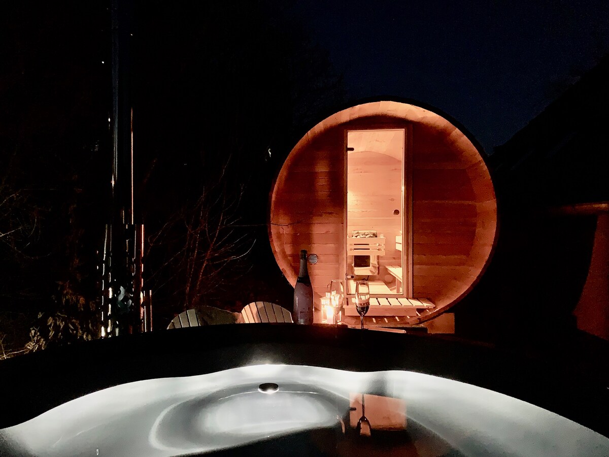 An illuminated wooden sauna stands beside a sleek hot tub, creating a serene ambiance at night. Soft reflections of light dance on the water's surface, while comfortable loungers are positioned nearby, inviting relaxation under the stars.