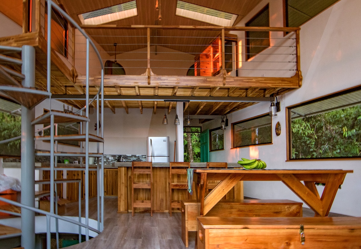 The main living area features an open layout with high ceilings and large windows, inviting natural light. The kitchen is equipped with wooden cabinetry, a refrigerator, and a dining table surrounded by benches. A wooden staircase leads to the mezzanine level above.