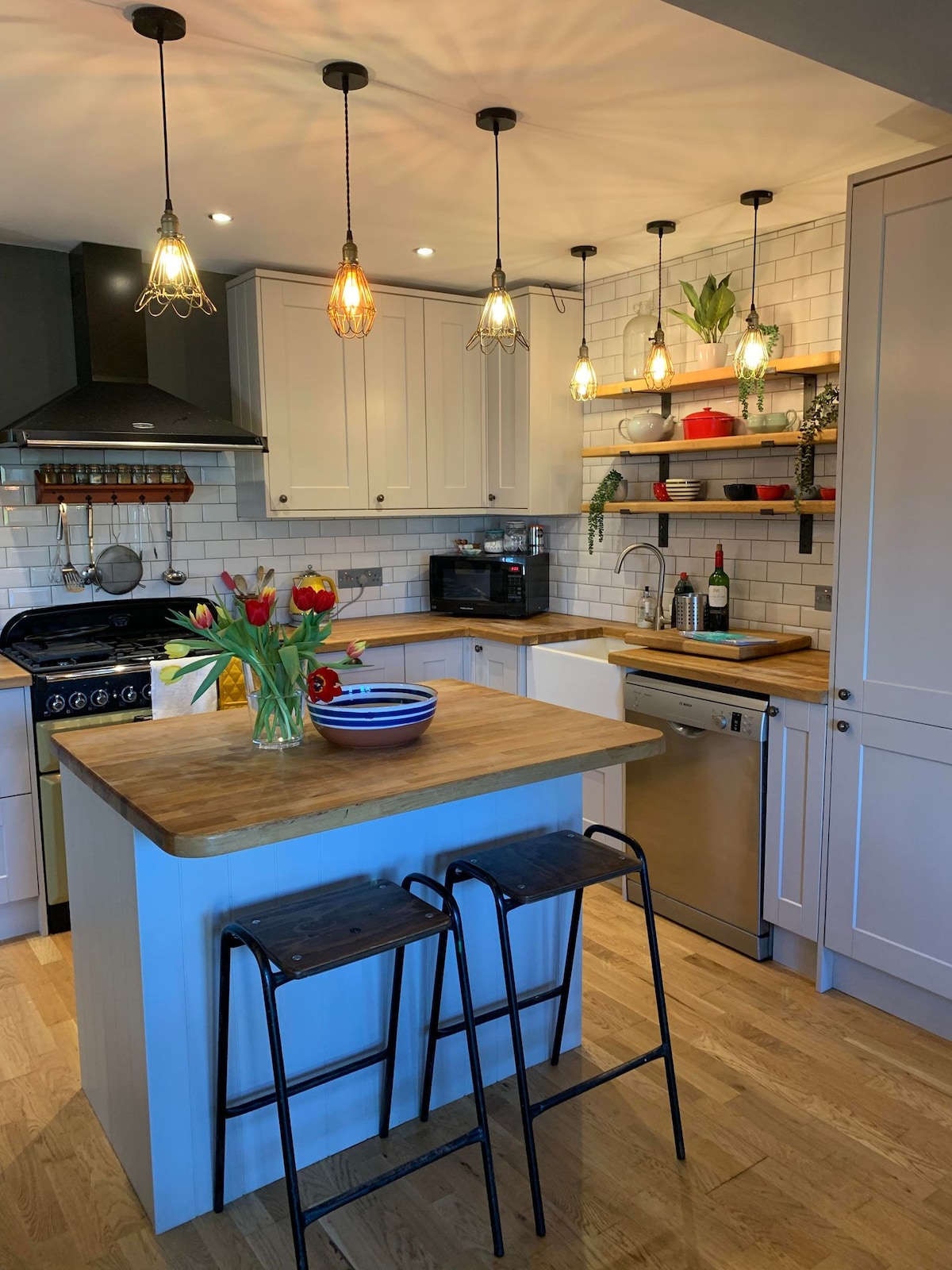 The kitchen features a central island with two stools, adorned with a vase of fresh flowers. Pendant lights hang above, illuminating the white cabinetry and tiled backsplash. Shelves display potted plants and kitchenware, while modern appliances are seamlessly integrated into the space.